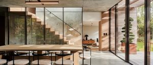 Dining room with floor to ceiling windows behind it. Staircase behind the windows and living room behind that. Floor to ceiling windows with views on the right.