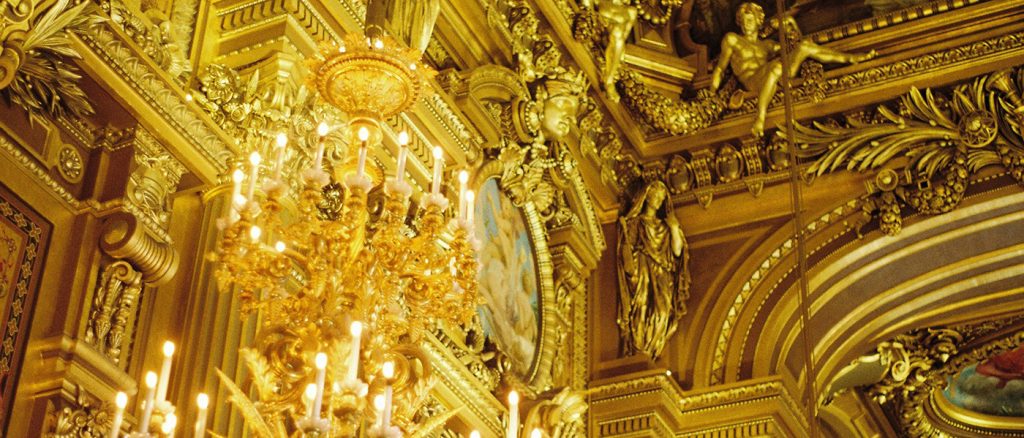 Gold chandelier, with gold cherubs on the roof of a gold ornate ceiling ad walls.